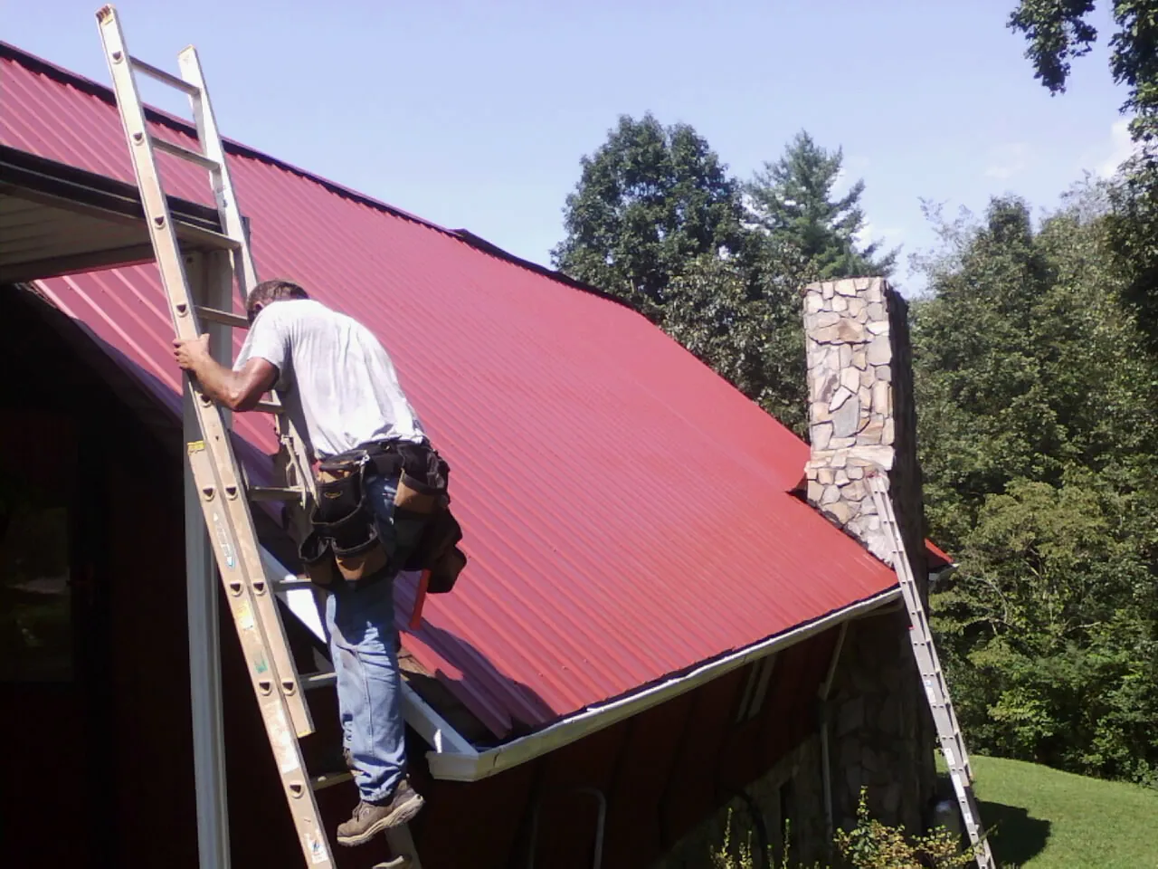 Roofer on a ladder beside a red metal roof and stone chimney — Soffit & Fascia Repair by Roofing Brothers in Nederland